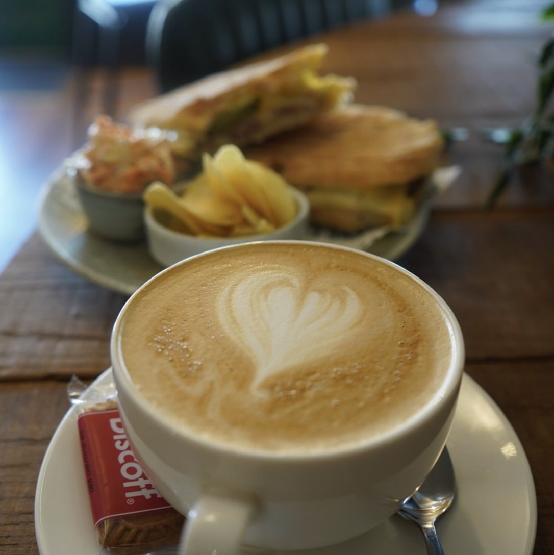 A flat white with latte art at The Little Deli, with a toastie behind it